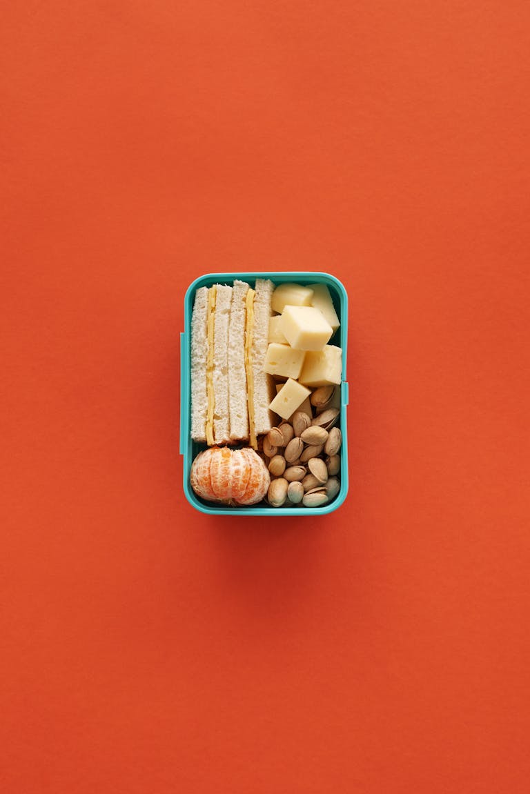 A balanced lunchbox featuring cheese, nuts, sandwich, and a tangerine against an orange background.
