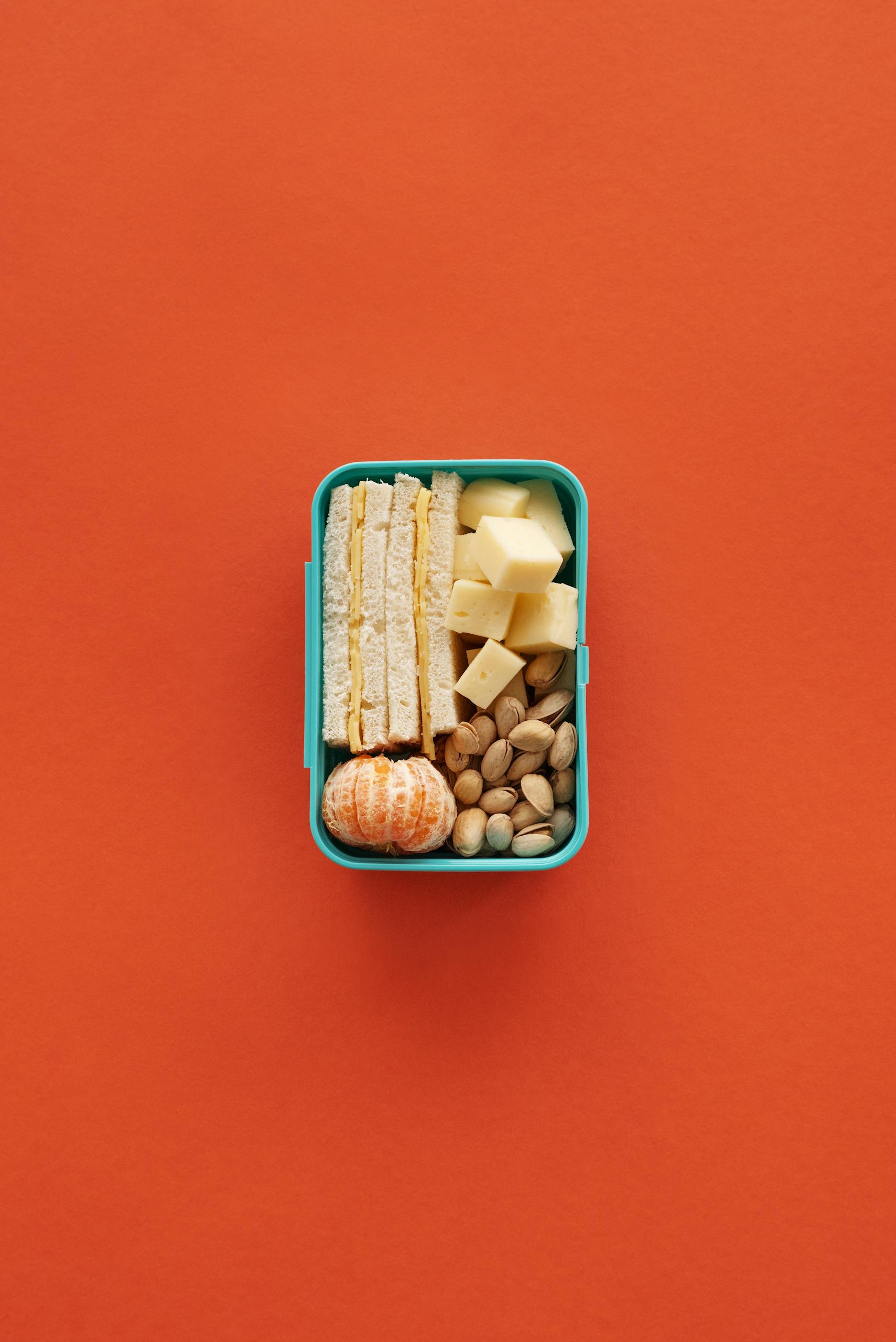 A balanced lunchbox featuring cheese, nuts, sandwich, and a tangerine against an orange background.