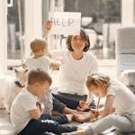 A mother holds a 'HELP' sign while sitting with her children and a dog in a sunlit living room.