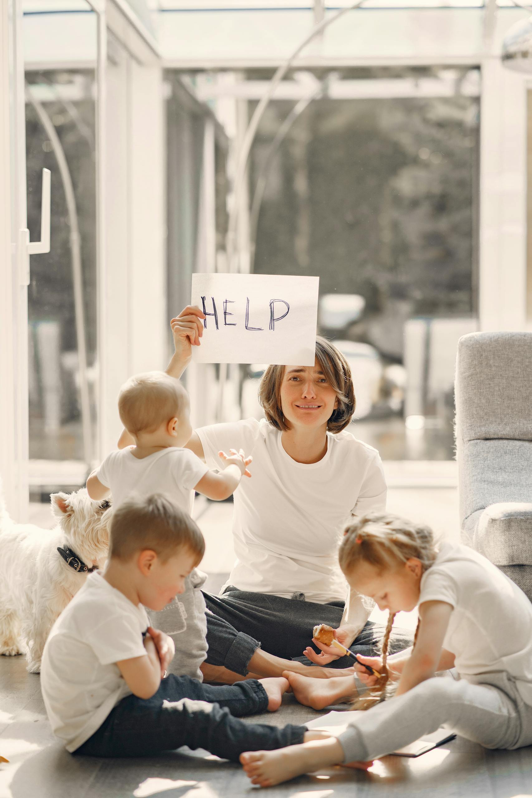 A mother holds a 'HELP' sign while sitting with her children and a dog in a sunlit living room.