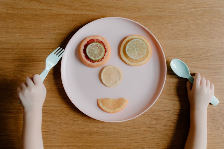 A playful citrus fruit face on a plate with child holding fork and spoon.
