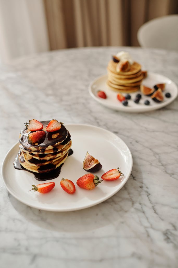 A tempting stack of pancakes topped with strawberries and figs, drizzled with chocolate syrup on a marble table.