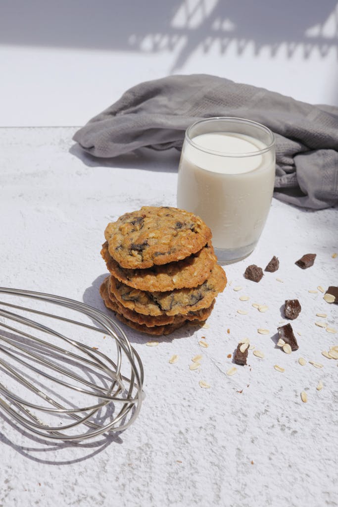 Delicious homemade cookies stacked beside a glass of milk and baking whisk on a bright day.