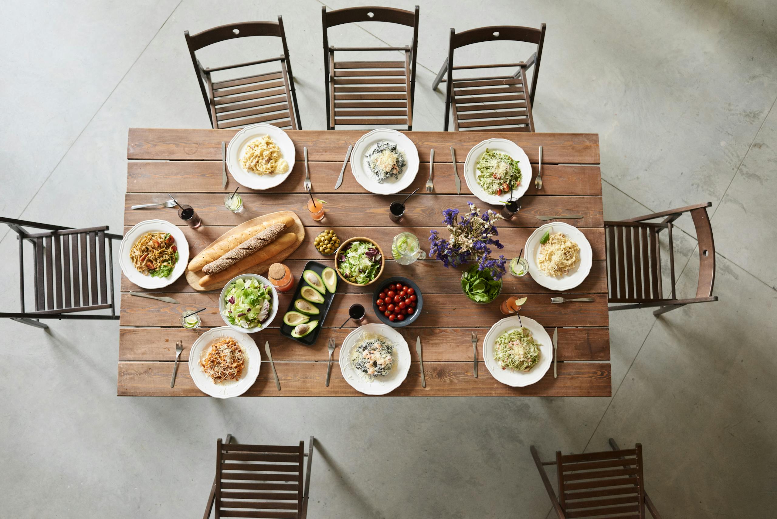 Top view of a wooden table set with pasta dishes, fresh ingredients, and drinks for a cozy meal.